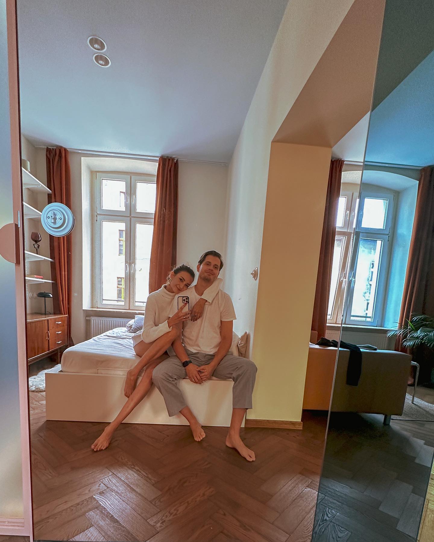Mirror selfie with Rafaela and her partner, sunlit Polish apartment, herringbone floors, orange linen curtains, full-length mirror.